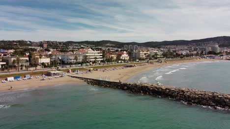 Sitges-coastline-with-city-buildings,-beaches,-and-clear-blue-water,-aerial-view