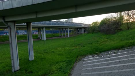 Drone-shot-of-car-on-elevated-bridge-with-green-grass-below-and-city-in-background