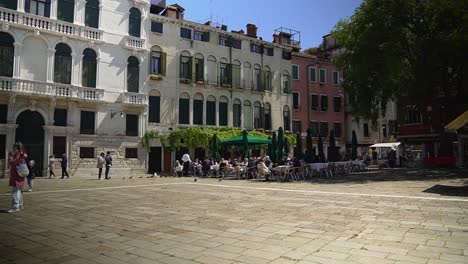 Toma-Panorámica-De-Una-Plaza-En-Venecia-Con-Gente-Sentada-Afuera-Del-Restaurante