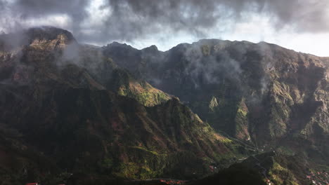 Low-clouds-moving-over-a-sunlit-mountainous-landscape,-in-Madeira---Aerial-view
