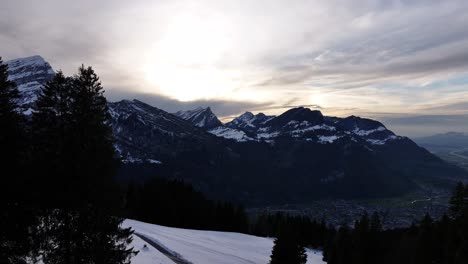 Vista-De-Los-Alpes-Suizos-Al-Atardecer,-Con-Laderas-Cubiertas-De-Nieve,-Siluetas-De-Pinos-Y-Majestuosos-Picos-Montañosos-Bajo-Un-Espectacular-Cielo-Nublado.