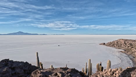 Pan-across-salt-flats-of-Bolivia-as-car-drives-below-cactus-covered-mountain