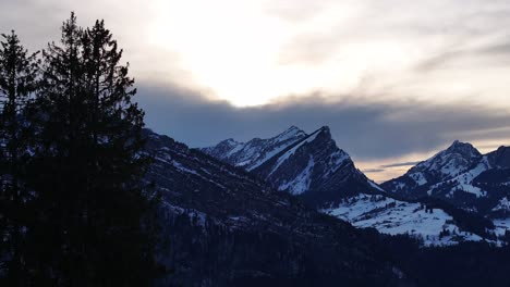 Aufschlussreiche-Aufnahme-Der-Schweizer-Alpen-Bei-Sonnenuntergang,-Mit-Kiefernsilhouetten-Im-Vordergrund-Und-Schneebedeckten-Berggipfeln-Im-Hintergrund-Unter-Einem-Dramatischen-Himmel