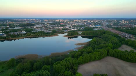 Panorama-at-sunrise,-drone-flight-over-the-forest-and-lake,-with-industrial-district-buildings-visible-in-the-background