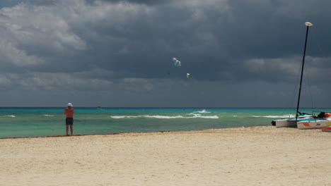 A-man-stands-on-a-beach-near-the-ocean-looking-out-at-paragliders-against-a-dark-dramatic-cloudy-sky