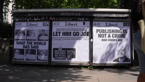 In-slow-motion-people-walk-past-posters-on-a-newsstand-depicting-newspaper-front-pages-with-the-headlines,-“What-the-world-has-said”,-“Let-him-go-Joe”-and,-“Publishing-is-not-a-crime,-war-crimes-are”