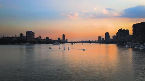 Aerial-perspective-above-the-surface-of-the-Nile-River-sunset-in-Downtown-Cairo,-Egypt,-boats-glide-gracefully-along-the-water,-embodying-the-concept-of-maritime-activity-and-urban-vitality