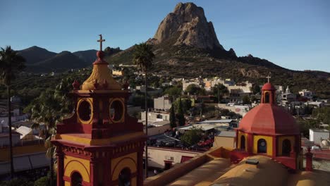 Red-and-yellow-bell-tower-of-a-famous-town-in-Mexico-called-Bernal,-with-one-of-the-three-largest-and-most-beautiful-monoliths-in-the-world-in-the-background,-a-sibling-of-the-Rock-of-Gibraltar