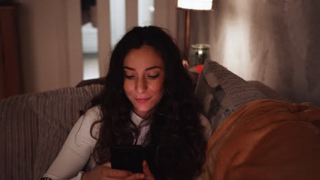 Young-Italian-woman-smiles-while-looking-at-her-smart-phone-at-home