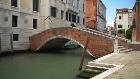 Scenic-small-bridge-crossing-a-canal-in-Venice-city,-two-people-standing-on-the-stone-bridge