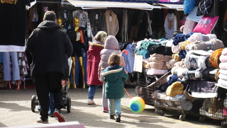 Los-Compradores,-Incluidos-Niños-Con-Globos-De-Colores,-Navegan-Por-Un-Mercado-Al-Aire-Libre-Lleno-De-Ropa-Y-Mantas.