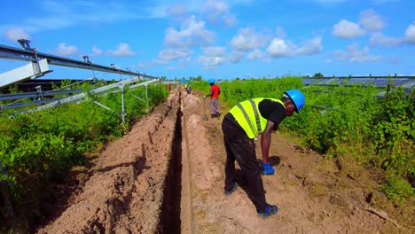 Trabajador-Africano-Con-Equipo-Completo-De-Ppe-Excavando-Zanjas-Para-La-Instalación-De-Cables-Eléctricos-En-Una-Granja-Solar