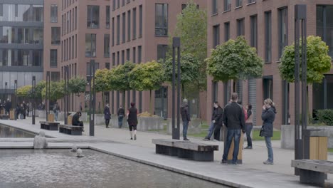 Pedestrians-stroll-by-modern-brick-buildings-along-a-landscaped-urban-waterway,-tilt-down-shot