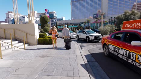 POV-Caminando-Detrás-De-Una-Pareja-Con-Equipaje-Rodante-A-Lo-Largo-De-La-Calle-Al-Lado-De-Una-Parada-De-Taxis-En-Las-Vegas-En-Un-Día-Soleado