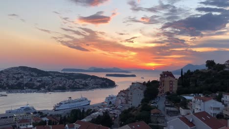 Time-lapse-of-dramatic-sunset-at-Dubrovnik-Port-Authority-in-Croatia-as-the-fiery-sun-dips-below-the-horizon,-illuminating-the-partially-cloudy-sky-with-bright-hues-of-orange-as-ships-pass