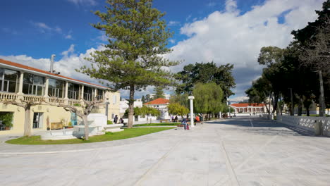 A-public-park-in-Pafos,-Cyprus,-with-a-two-story-building,-trees,-and-people-enjoying-the-sunny-day