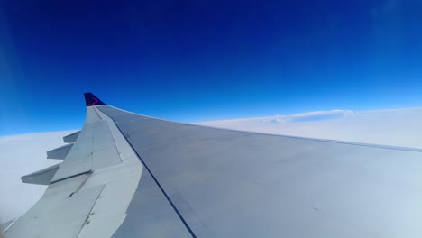 View-of-airplane-wing-with-beautiful-snowy-white-clouds-and-deep-blue-sky-on-sunny-day-shot-through-aircraft-window