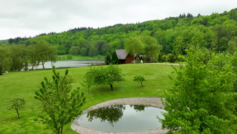 A-serene-park-landscape-with-a-small-pond,-trees,-and-a-cozy-cabin-in-the-background,-surrounded-by-green-hills