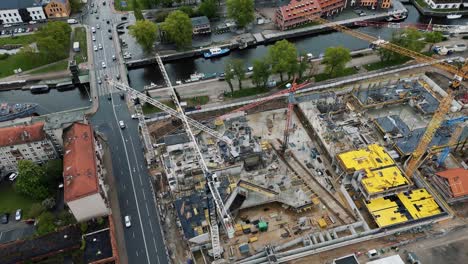 A-large-urban-construction-site-by-a-river-with-cranes-and-busy-roads-nearby,-aerial-view