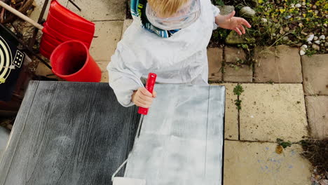 Un-Niño,-Equipado-Con-Gafas-Protectoras,-Utiliza-Un-Rodillo-Para-Pintar-Una-Mesa-Al-Aire-Libre