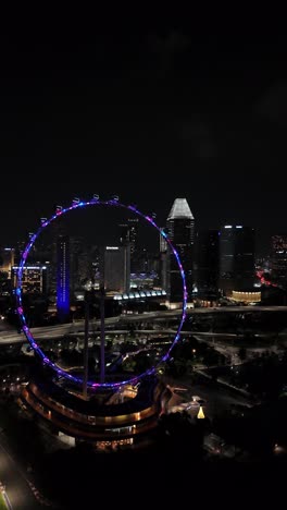 Rotating-nighttime-aerial-of-the-beautifully-lit-up-Singapore-Flyer-ferris-wheel-in-the-Marina-Bay-Sands-area,-vertical-aerial-view