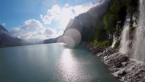 Aerial-flight-next-to-a-beautiful-big-waterfall-on-a-mountain-Landscape,-Drone-Flying-Over-a-Blue-Lake---Oeschinen-Lake,-Switzerland