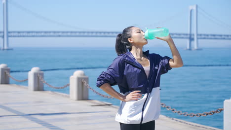 Attractive-sportswear-sporty-woman-drinking-water-after-workout-jogging-fitness-training-in-activewear-standing-on-sea-bank-near-bridge-view