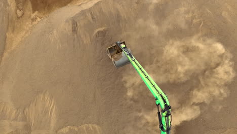 An-aerial-shot-of-an-excavator-digging-into-a-large-sand-pile,-lifting-sand-and-creating-a-dust-cloud