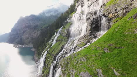 Luftflug-Neben-Einem-Wunderschönen-Großen-Wasserfall-In-Einer-Berglandschaft,-Drohne-Fliegt-über-Einen-Blauen-See---Oeschinensee,-Schweiz