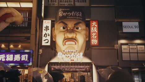 Gente-Con-Sombrillas-Frente-Al-Restaurante-Kushikatsu-Daruma-dotombori-En-Una-Noche-Lluviosa-En-Osaka,-Japón