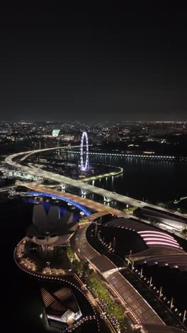 Atemberaubende-Nächtliche-Luftaufnahme-Des-Beleuchteten-Marina-Bay-Sands-Gebiets-Mit-Dem-Berühmten-Riesenrad-Singapore-Flyer,-Beleuchtet-Am-Nachthimmel,-Vertikales-Luftbildvideo