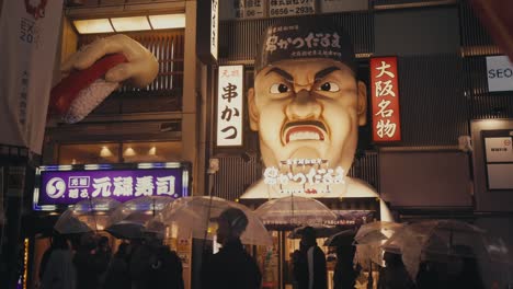 People-Walking-In-Front-Of-Kushikatsu-Daruma-At-Dotonbori-Street-On-A-Rainy-Night-In-Osaka,-Japan