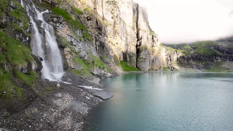 Luftflug-Neben-Einem-Wunderschönen-Großen-Wasserfall-In-Einer-Berglandschaft,-Drohne-Fliegt-über-Einen-Blauen-See---Oeschinensee,-Schweiz