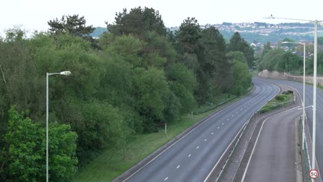 Leere-Zweispurige-Straße-In-Den-Frühen-Morgenstunden-Mit-Häusern-Auf-Mit-Bäumen-Bewachsenen-Hügeln-Im-Hintergrund