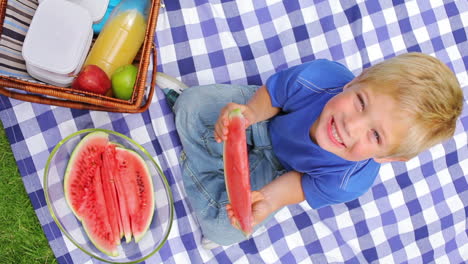 Overhead-shot-of-a-boy-sitting-on-a-picnic-blanket-while-eating-a-slice-of-watermelon-and-smiling