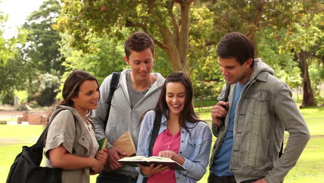 Four-friends-laughing-while-pointing-at-a-book-as-they-stand-together-in-a-park