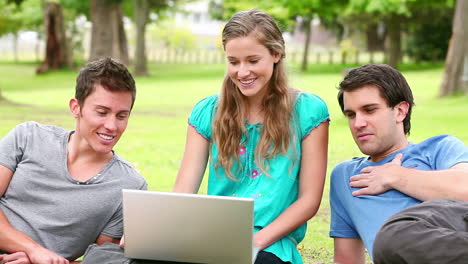 Woman-laughing-and-talking-as-she-uses-a-laptop-while-sitting-with-her-two-friends-in-a-park