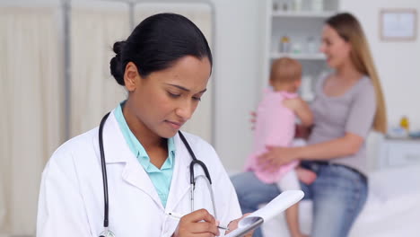 Doctor-writing-on-a-clipboard-with-a-woman-and-her-baby-in-background