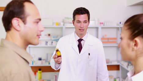 Smiling-pharmacist-pointing-at-a-flask-of-pills-to-a-man-and-a-woman