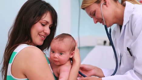 Mother-holding-a-baby-in-an-examination-room-with-a-smiling-doctor-using-a-stethoscope