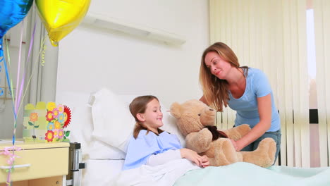 Smiling-woman-giving-a-teddy-bear-to-a-smiling-girl-in-a-bed