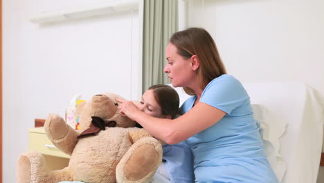 Woman-giving-a-teddy-bear-to-a-girl-in-a-bed-with-balloons