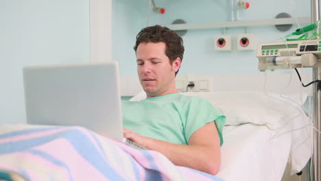 Smiling-male-patient-typing-on-a-laptop-while-lying-on-a-bed