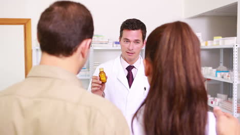 Pharmacist-holding-at-a-flask-of-pills-to-a-man-and-a-woman