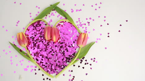 Luz-Brillando-Sobre-Un-Corazón-Hecho-De-Confeti-Rosa-Enmarcado-Por-Tulipanes