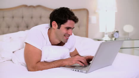 Hombre-Guapo-Sonriente-Usando-Una-Computadora-Portátil-En-La-Cama