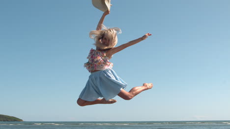 Attractive-blonde-holding-straw-hat-jumping-on-the-beach