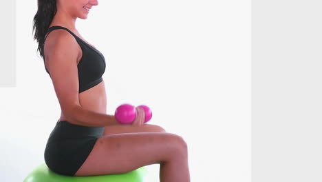 Side-view-of-fit-woman-sitting-on-exercise-ball-training-with-dumbbells
