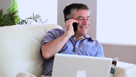 Happy-man-talking-on-the-phone-on-the-sofa-with-his-laptop