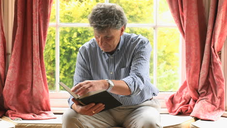 Focused-mature-man-sitting-by-a-window-reading-a-book